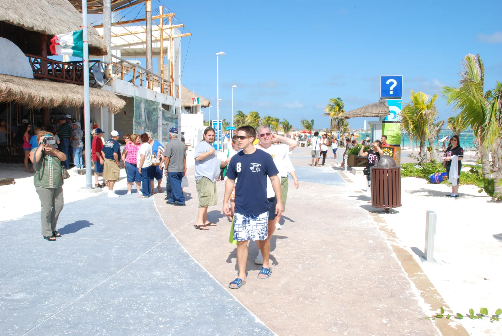 Pedestrians on Mahahual’s beachfront malecón beside waterfront buildings and palm trees